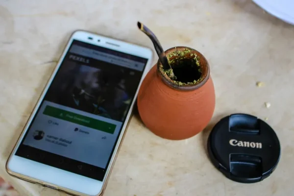 A flat lay of a smartphone, yerba mate cup, and camera lens cap on a table.