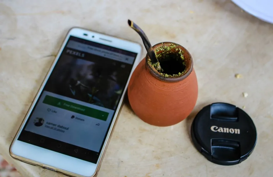 A flat lay of a smartphone, yerba mate cup, and camera lens cap on a table.
