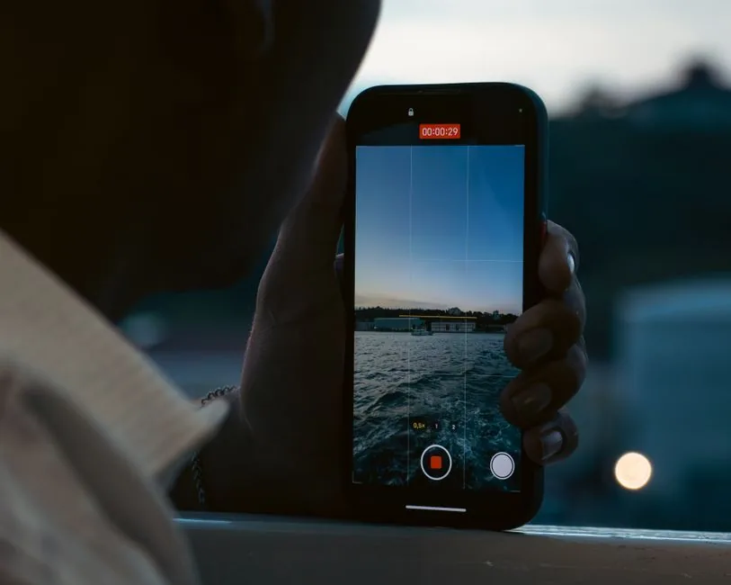 A person records a peaceful waterfront scene using a smartphone at dusk.