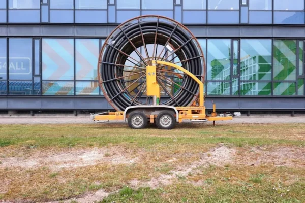 A large industrial cable reel on a trailer parked outside a modern glass building in Amsterdam.